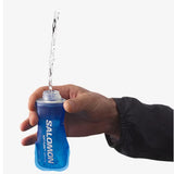 Hand holding a blue Salomon water bottle with water being poured from it on a white background